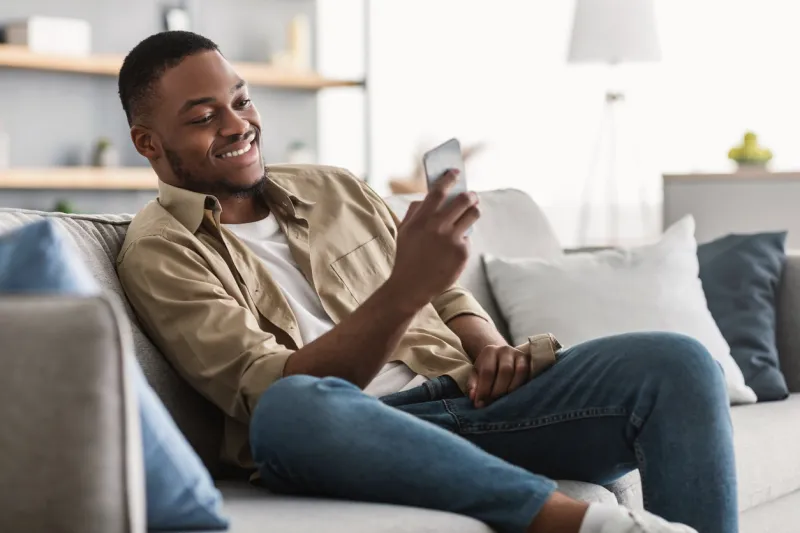 side view of african american guy using smartphone browsing internet sitting on couch at home black man texting on cellphone or using new application on mobile phone indoors