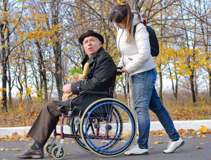 woman pushing an elderly handicapped man in a wheelchair along a rural street as they return from doing the grocery shopping together