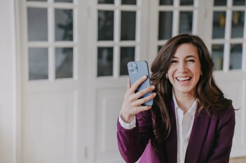 excited brunette woman in violet suit toothy smiling looks at camera holding phone happily, received great news at office successful brunette young adult businesswoman satisfied by new smartphone