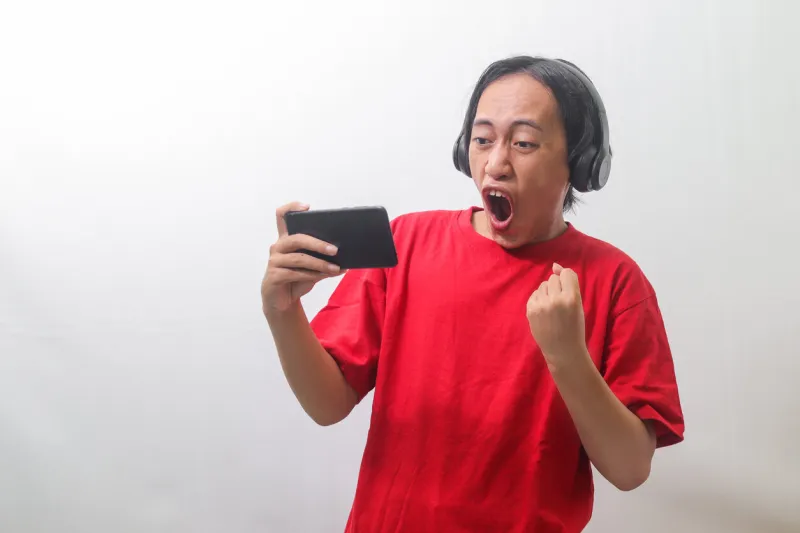 portrait of excited asian man in red t-shirt raising his fist, celebrating winning game on his mobile phone isolated image on gray background