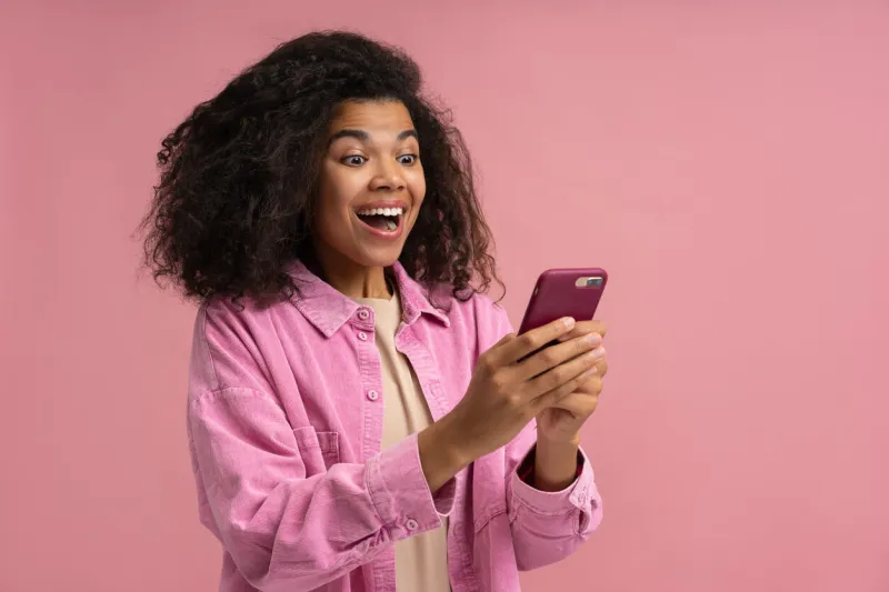 portrait of excited african american woman holding smartphone playing mobile game isolated on pink background emotional hipster using mobile phone shopping online with big sales innovation, modern technology