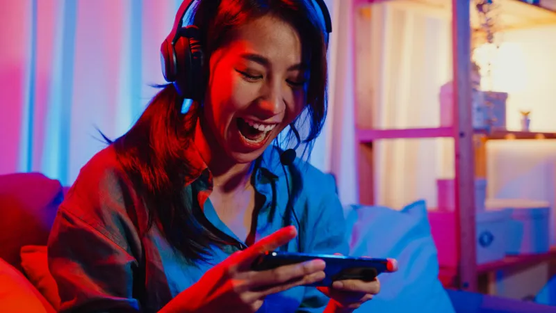 happy asia girl gamer wear headphone competition video game online with smartphone excited talk with friend sit on couch in colorful neon lights living room at home home quarantine activity concept