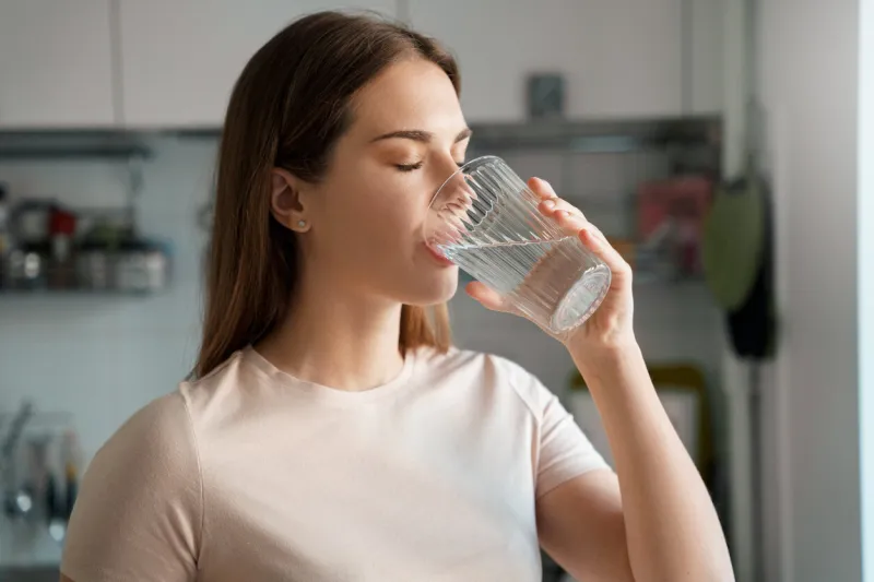 thirsty young woman drinking fresh water from glass home office kitchen interior headshot portrait dehydration prevention, normal bowel function and balance of body maintenance concept