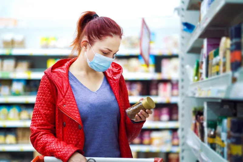 red haired woman in red jacket wearing medical mask choosing jar of instant coffee at grocery store and putting it in shopping cart, blurred people passing by on background concept of pandemic