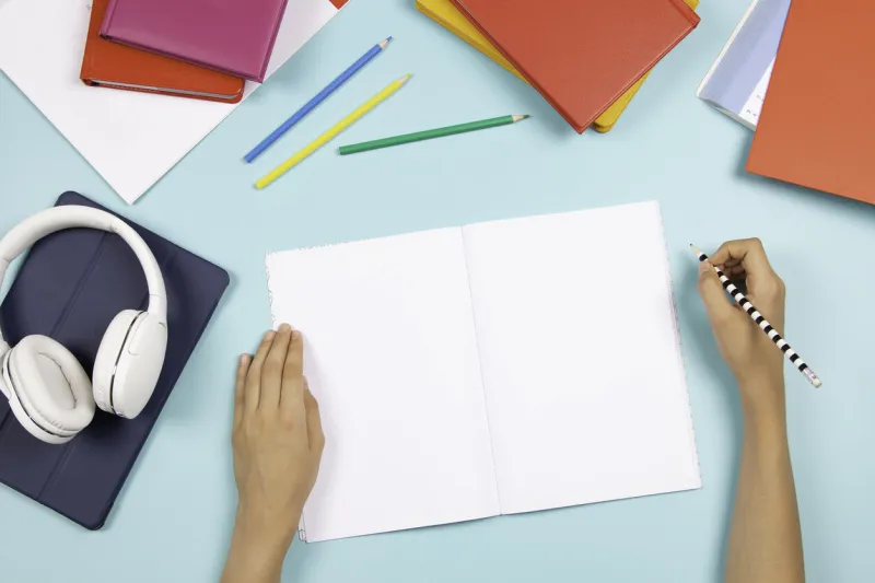 kid hand holding pencil and writing in blank open notebook digital tablet, headphones, books, school stationary on blue background back to school, language learning, online classes, homeschooling