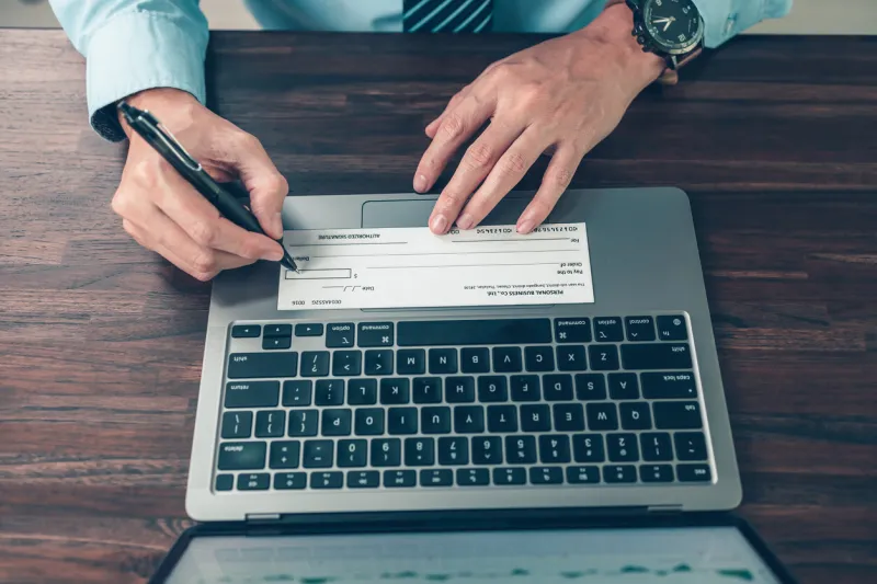 business man hand writing and signing white blank bank check book and dollar bill, coin, laptop and graph chart on the desk at office payment by check, paycheck, payroll concept