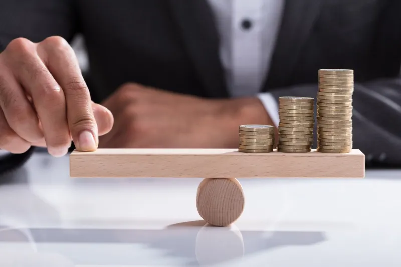 close-up of a businessperson's hand balancing stacked coins on wooden seesaw with finger over desk