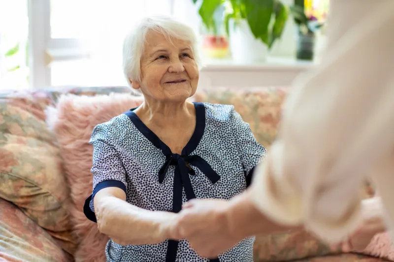 female home carer supporting old woman to stand up from the sofa at care home