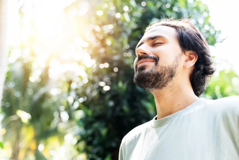 a bearded man is meditating outdoor in the park with face raised up to sky and eyes closed on sunny summer day concept of meditation, dreaming, wellbeing and healthy lifestyle