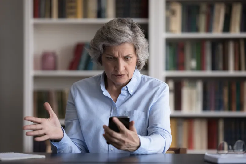 angry nervous middle aged woman looking at cellphone screen, feeling anxious unhappy getting message with bad news, dissatisfied with bad work or broken device in office, sitting alone at table