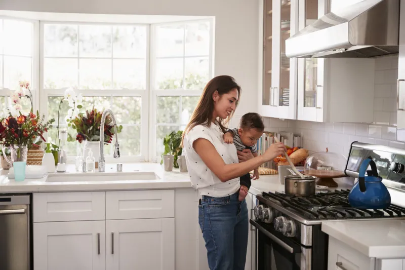 multitasking mum holding her young baby while she makes food at the hob in her kitchen, side view
