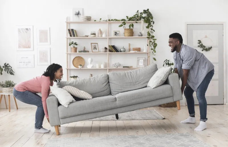 happy african american couple moving sofa in living room, replacing furniture at home, side view