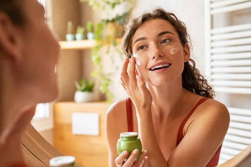 woman caring of her beautiful skin face standing near mirror in the bathroom young woman applying moisturizing cream on her face during morning routine smiling natural girl holding little green jar of ecological cosmetic cream