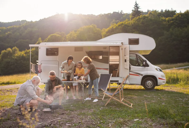 a multi-generation family sitting and eating outdoors by car, caravan holiday trip