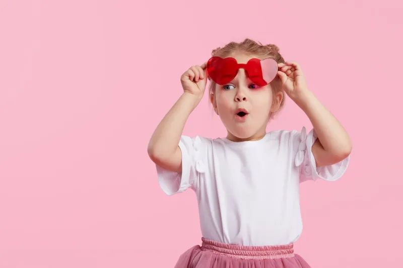 portrait of surprised cute little toddler girl in the heart shape sunglasses child with open mouth having fun isolated over pink background looking at camera wow funny face