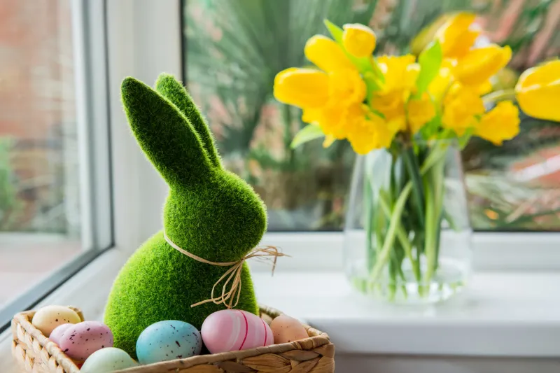 easter bunny rabbit statuette in straw basket with colored eggs on the windowsill with fresh spring tulips and daffodils flowers bouquet on the background happy easter selective focus copy space