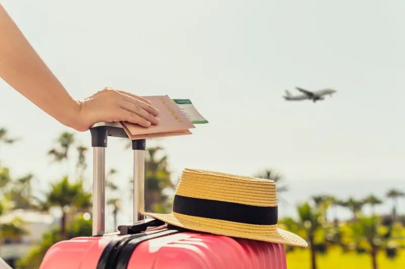 woman with pink suitcase and passport with boarding pass standing on passengers ladder of airplane opposite sea with palm trees tourism concept