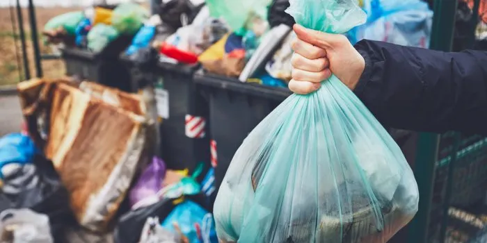hand with garbage against full trash cans with rubbish bags overflowing onto the pavement