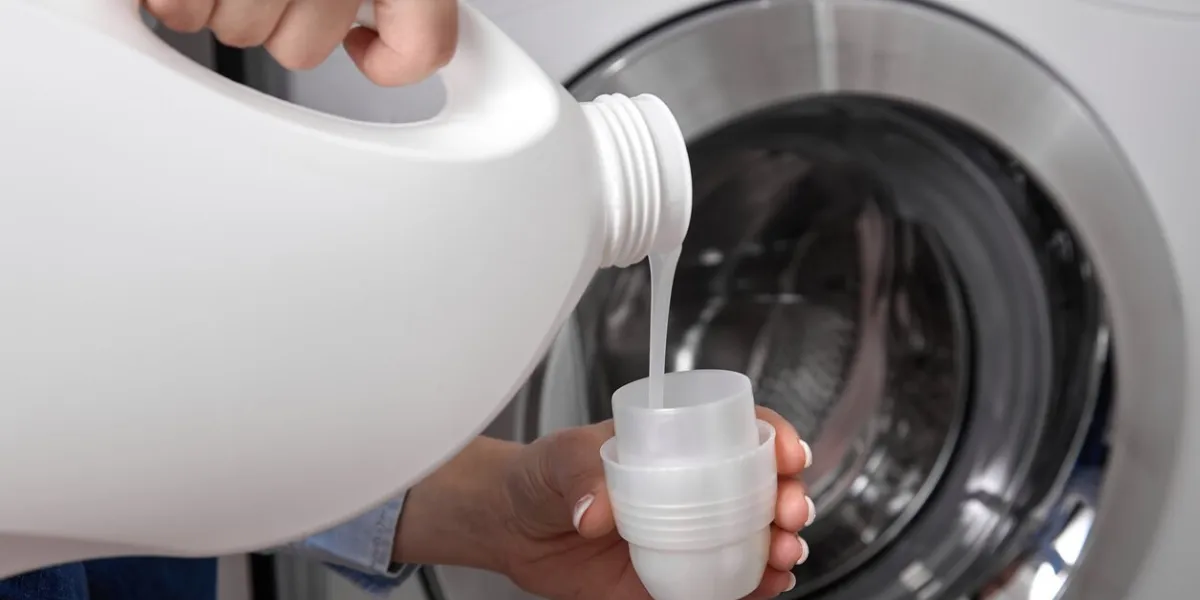against background of drum of steel-colored washing machine, woman pours liquid washing gel into plastic cap a girl in a white t-shirt carefully pours a transparent conditioner for flattening laundry