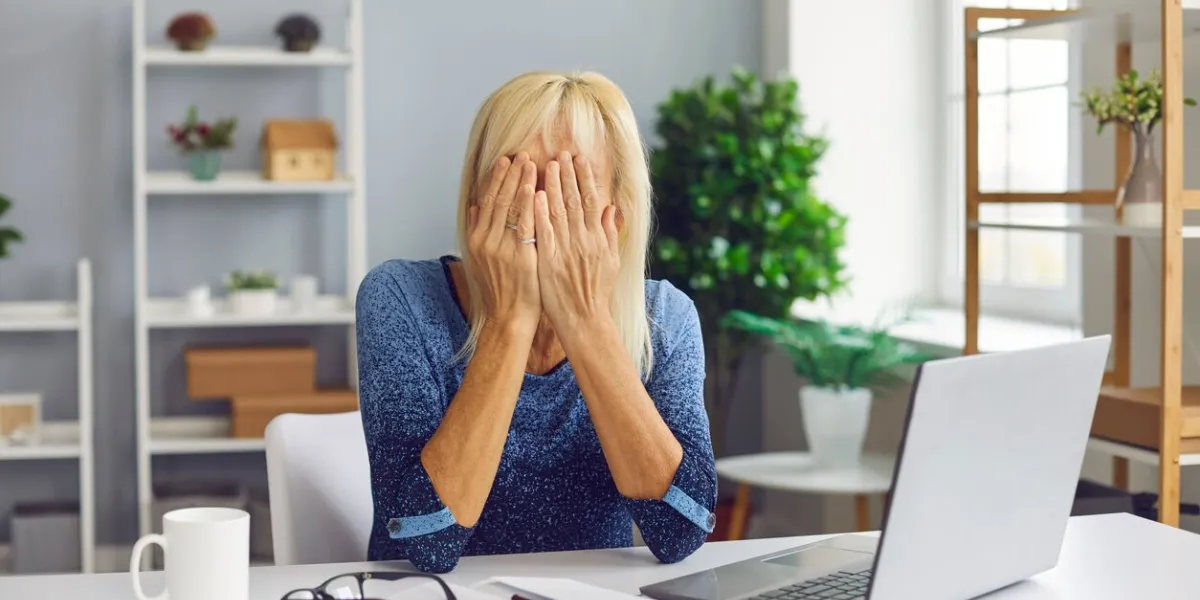 frustrated senior woman covering face unable to deal with problem online fraud victim scammed on internet crying, sitting at desk with laptop computer stressed mature lady having financial trouble