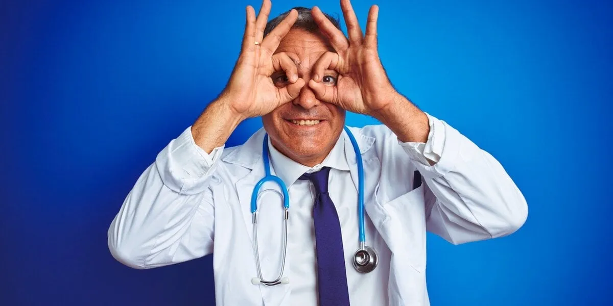 handsome middle age doctor man wearing stethoscope over isolated blue background doing ok gesture like binoculars sticking tongue out, eyes looking through fingers crazy expression