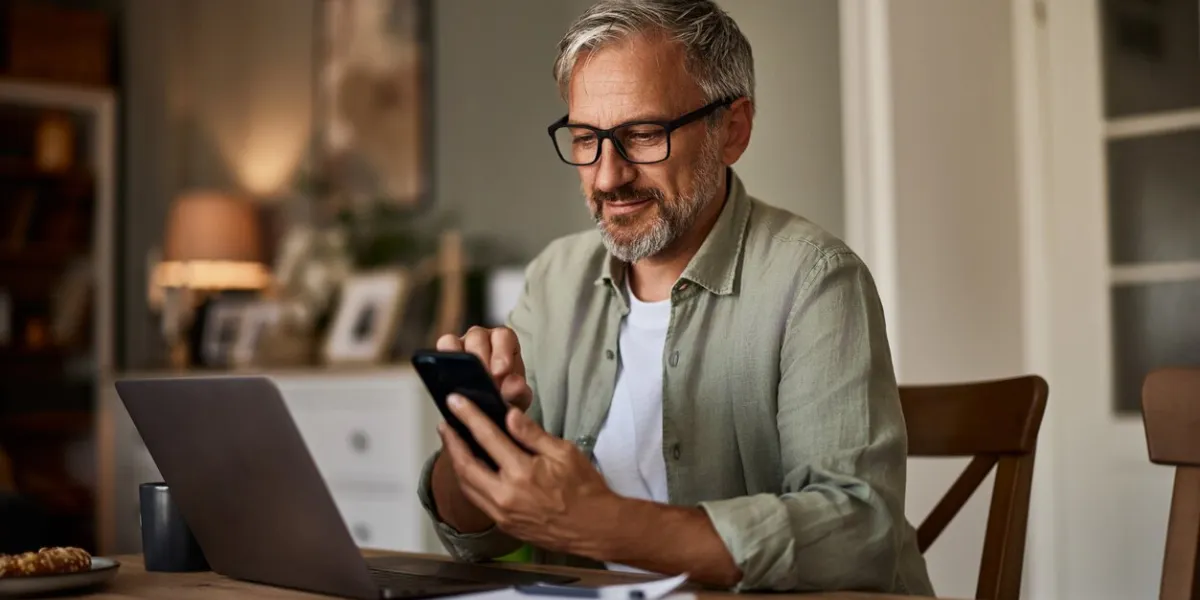 a man working from home over a laptop and using a mobile phone sitting at the wooden table