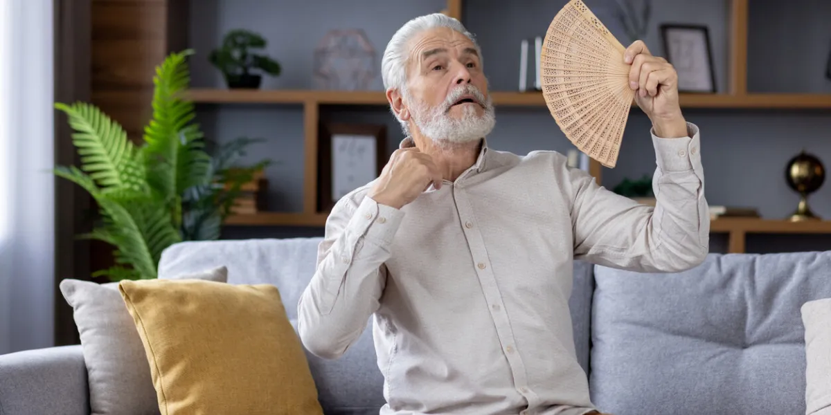 elderly caucasian man sitting on a gray sofa, using a handheld fan to cool off during a warm day indoors, looking uncomfortable and sweaty in a well-decorated living room