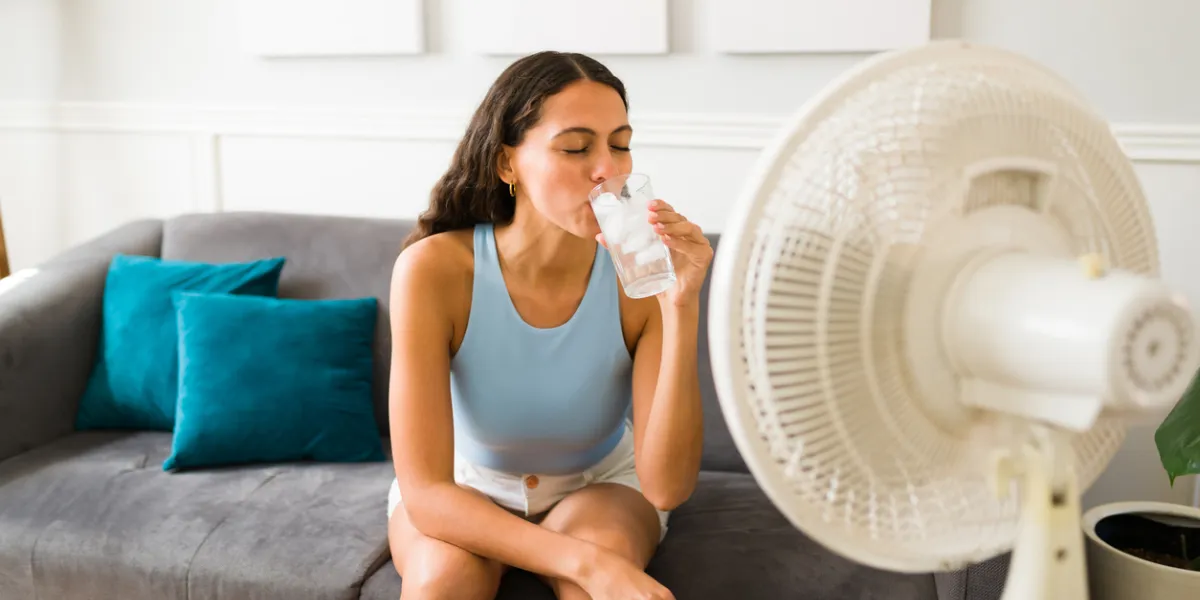 Une femme boit un verre d'eau frais devant un ventilateur