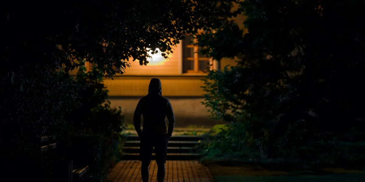 hooded man standing on sidewalk at dark park in summer black night scary moment and gloomy atmosphere thief watching house back view