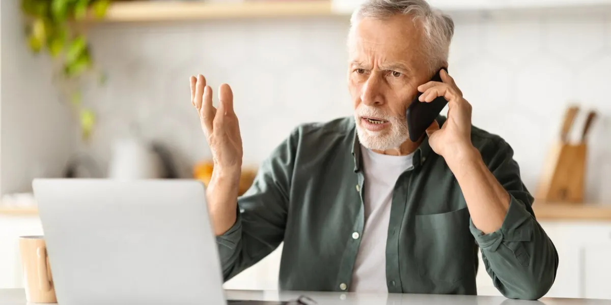 stressed senior man talking on cellphone and using laptop in kitchen, anxious elderly gentleman speaking with customer support, having problems with computer or internet, closeup shot