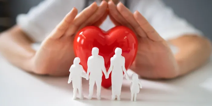 doctor's hand protecting red heart with family figure on reflective desk