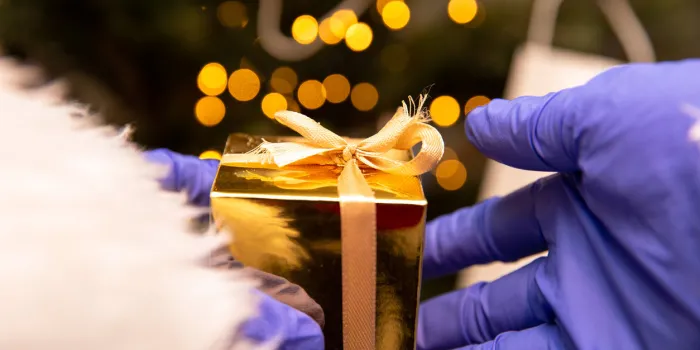 santa claus with gloves and face mask for coronavirus by the christmas tree, holding a golden present, covid-19 and christmas safety concept closeup