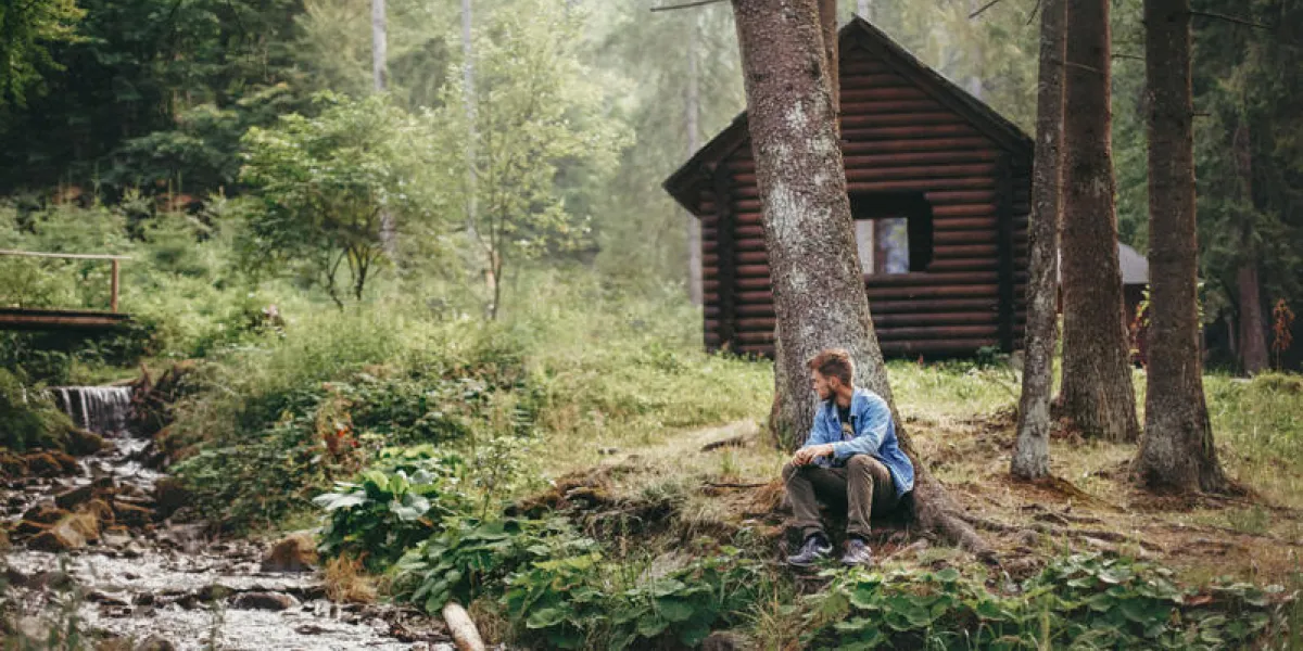 stylish hipster man sitting and relaxing at wooden cabin in forest guy exploring woods and river space for text travel and wanderlust concept