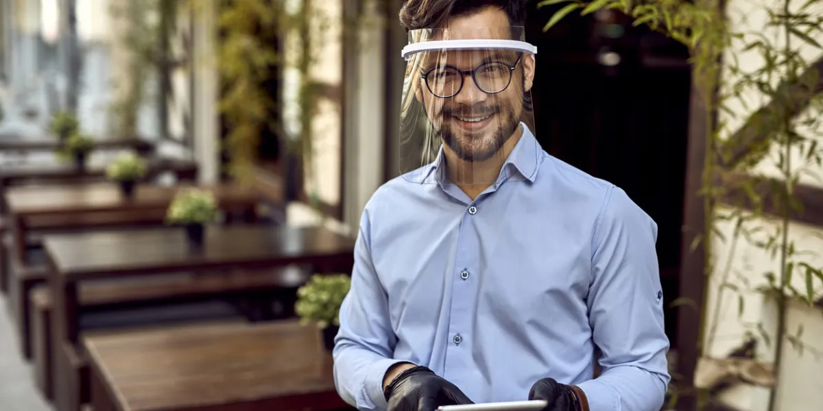 happy waiter wearing visor while working on touchpad at outdoor cafe and looking at camera