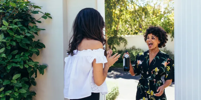 excited african woman attending a female friend's housewarming party woman giving a wine bottle her friends at entrance door