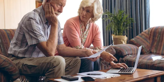front view of sad caucasian senior couple discussing bill while sitting on vintage sofa at retirement home senior woman is using laptop