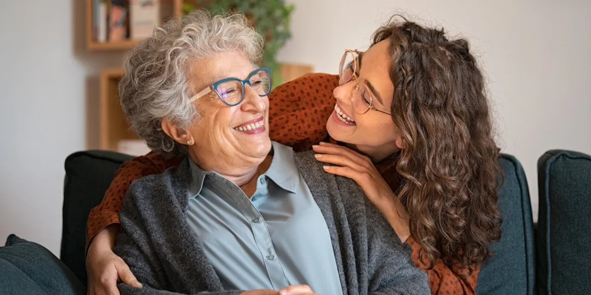 old grandmother and adult granddaughter hugging at home and looking at each other happy senior mother and young daughter embracing with love on sofa happy young woman hugging from behind grandma with love