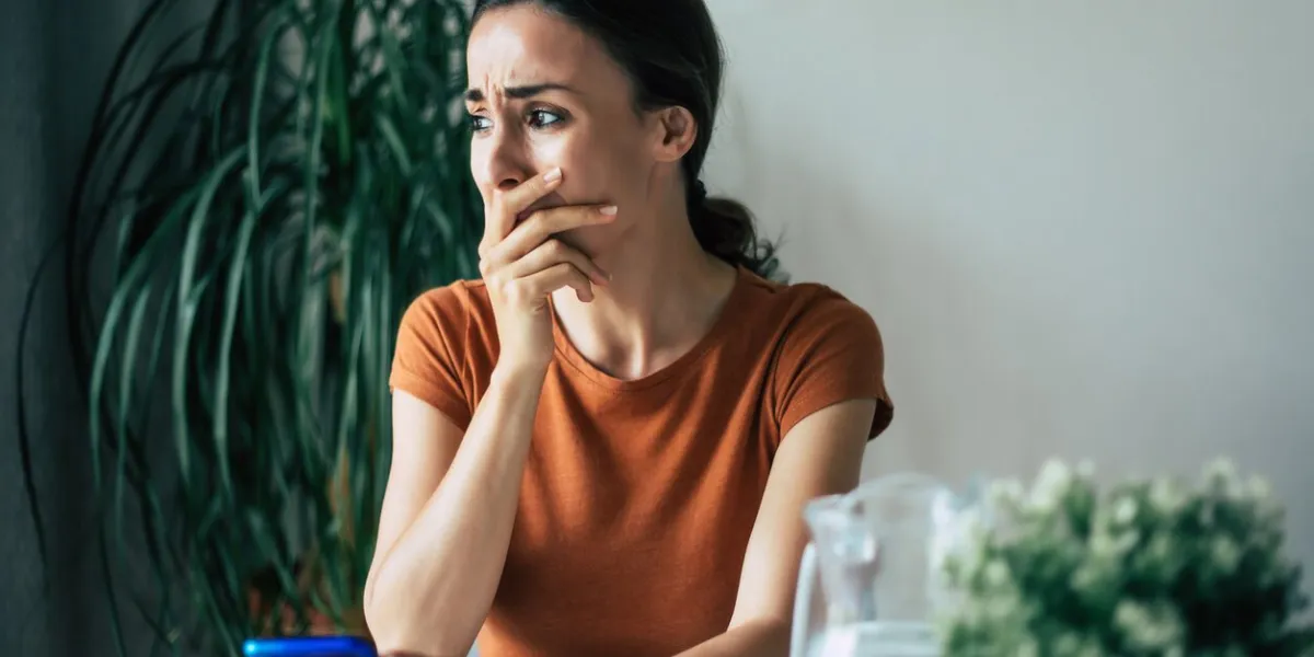 sad, frustrated young brunette woman is crying with smartphone in hands while she sitting on the chair at apartment