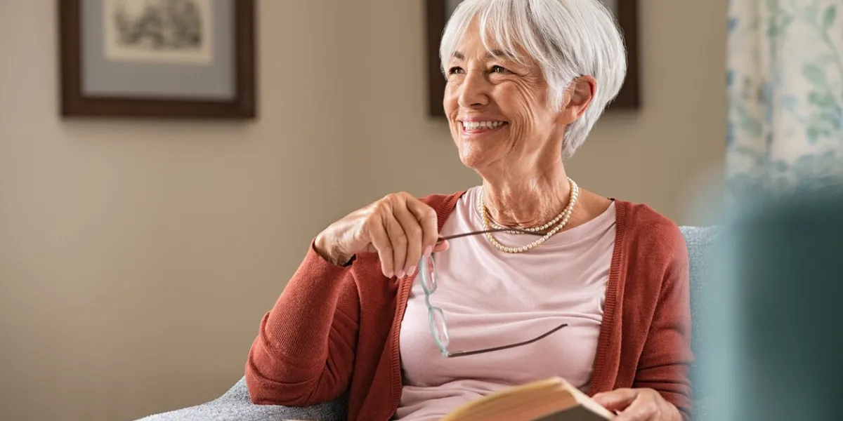cheerful senior woman holding book and eyeglasses thinking while relaxing at home happy elderly woman reading book at home sitting on couch beautiful old teacher takes a break from reading while looking through the window with a big grin
