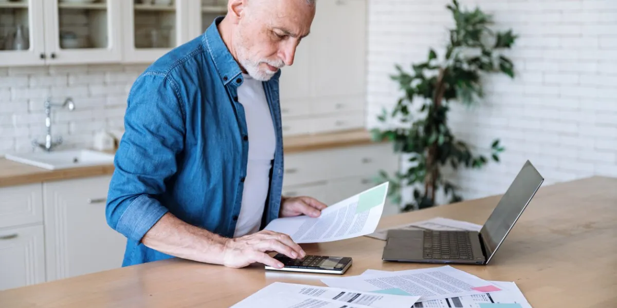 focused middle aged man making calculations of utility payment holding paper bill invoice in hand at table serious businessman doing paperwork for paying taxes online using laptop at home