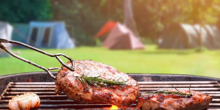roasted beef ripe steaks on flaming charcoal grill with blurred background of camping area in natural parkland