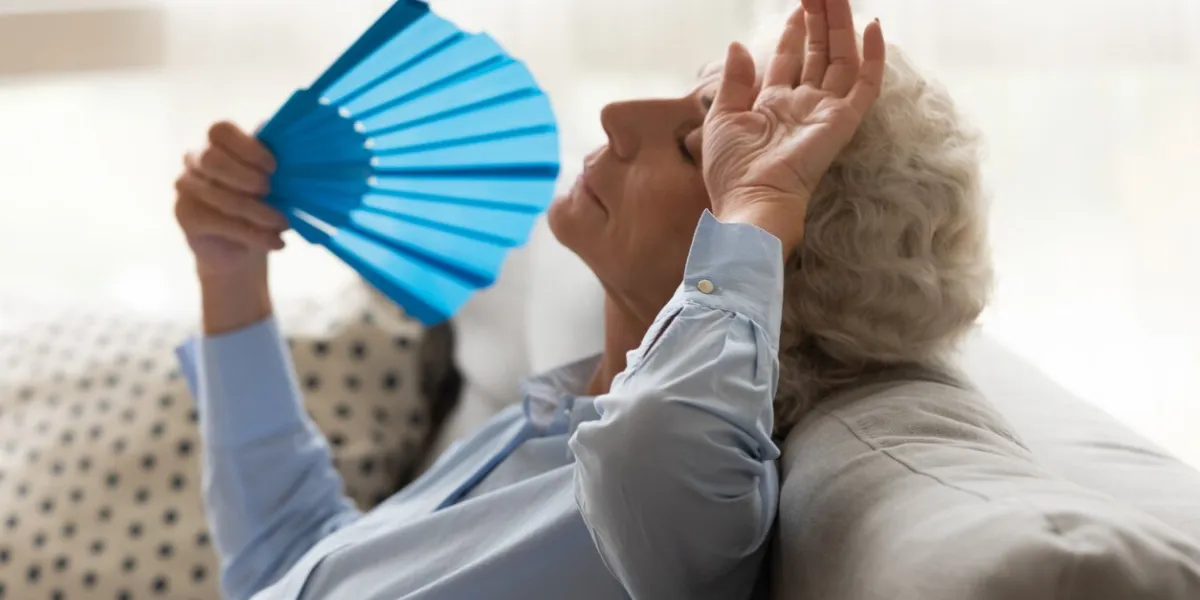 exhausted older woman feeling unwell, touching forehead with hand, suffering from heat, waving blue paper fan close up, sitting on couch, mature female cooling in hot summer weather, high temperature