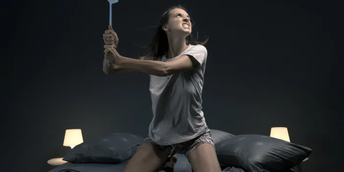 angry woman killing mosquitoes in her bedroom, she is sitting on the bed and holding a fly swatter like a weapon