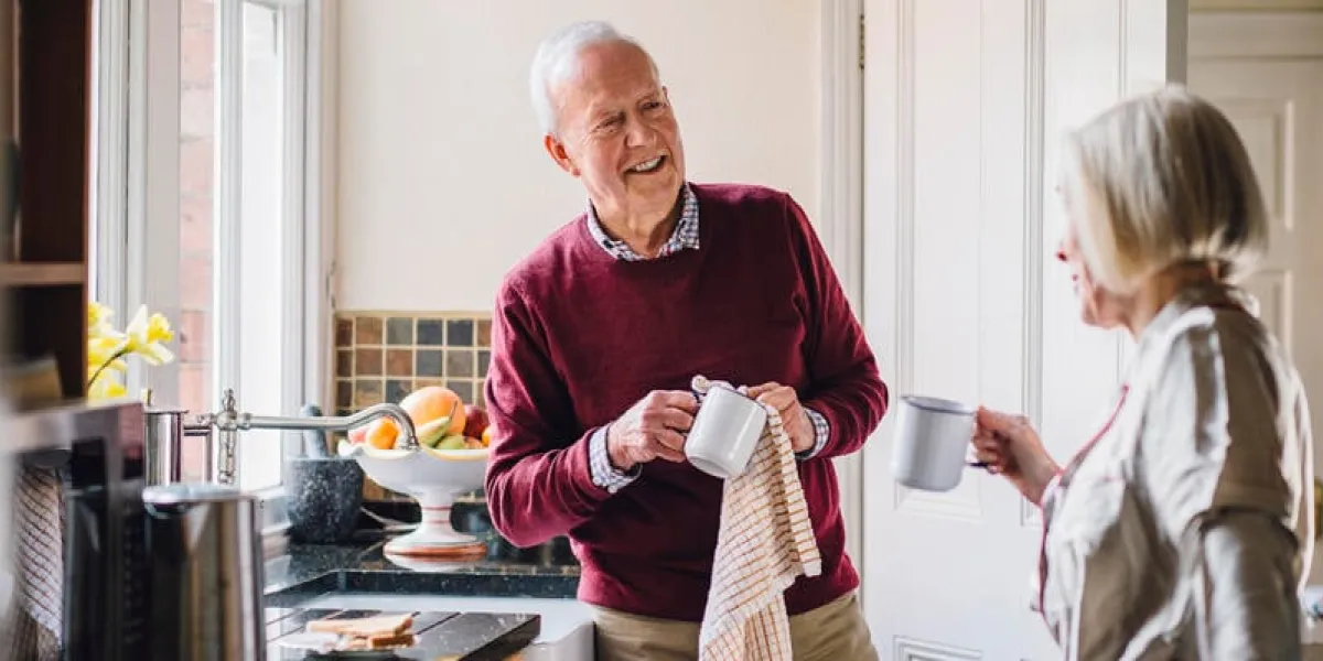 senior man is talking to his wife in the kitchen he is drying dishes and the woman is dirnking tea