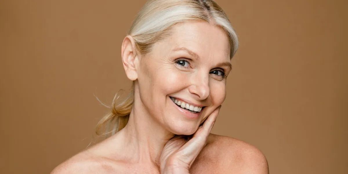 pretty charming woman with blonde hair looks seriously at camera touches face and enjoys softness of skin after spa procedures wears casual top isolated over brown background natural beauty portrait