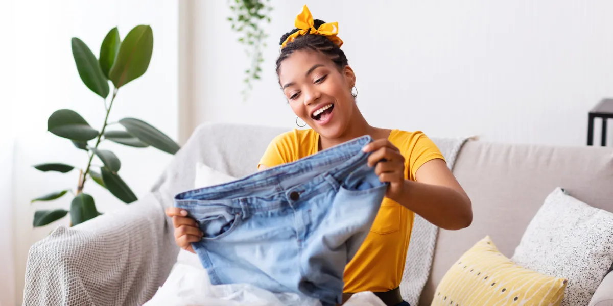 delivery concept happy black woman sitting on couch and holding jeans, unboxing cardboard package, free space