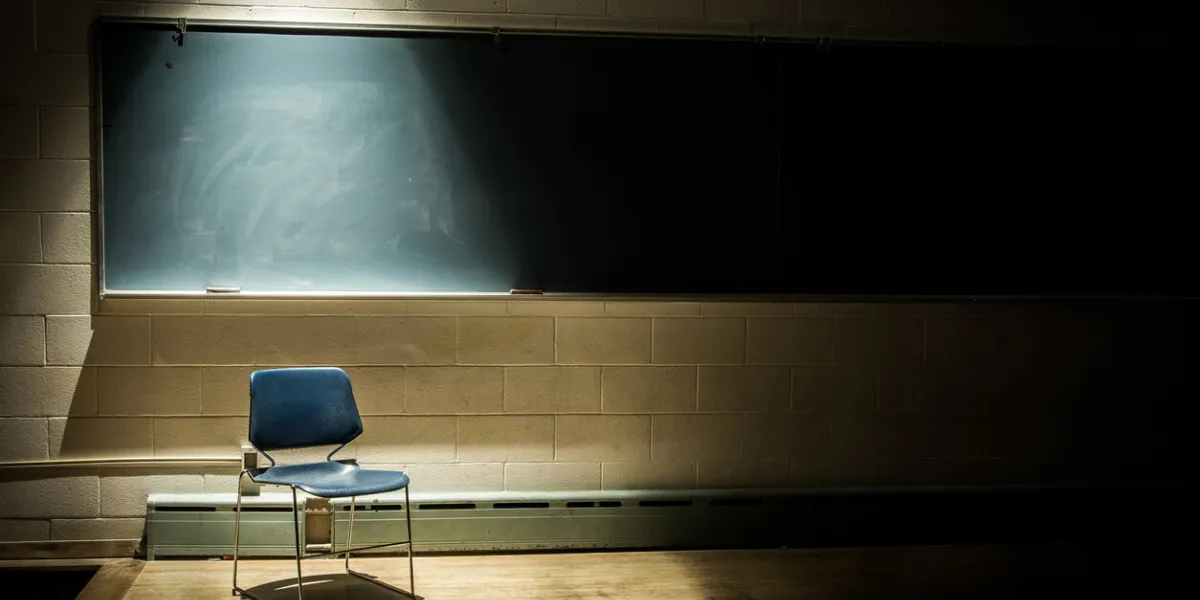 an empty school chair in a dark, shadowy classroom - in front of a chalkboard with a single beam of light overhead