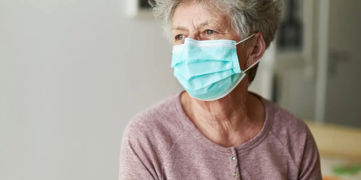 a senior citizen sits alone on her bed with a respirator or surgical mask and looks sadly and frightened out of the window and into the camera