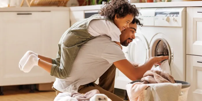 side view of black child in casual clothes with curly hair smiling and embracing dad loading washing machine during household routine in morning at home