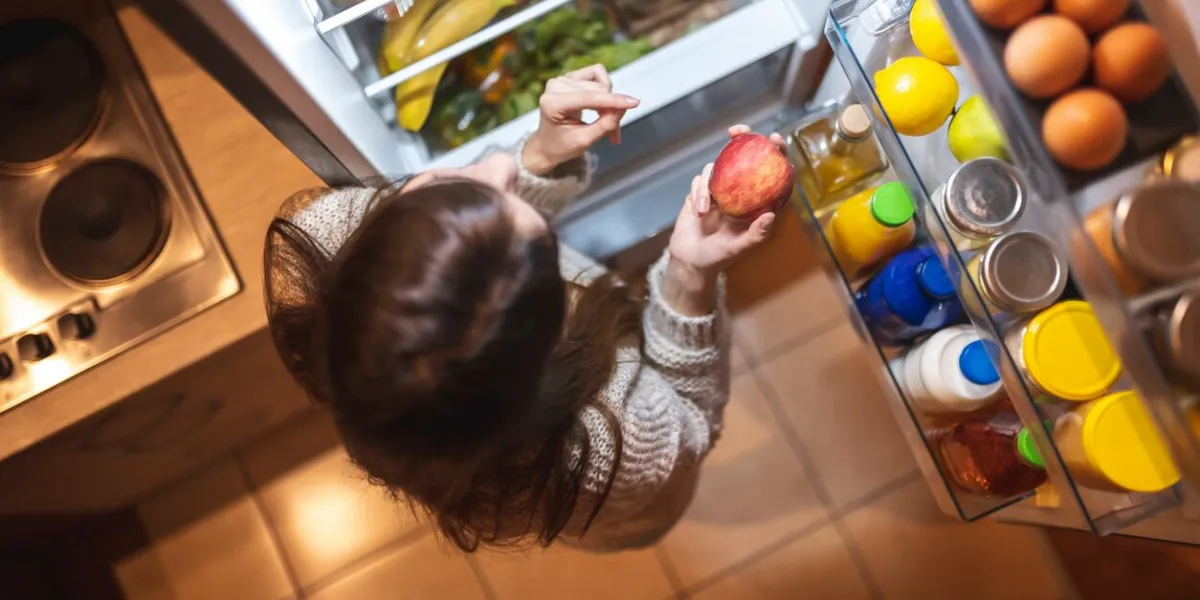 top view of a beautiful young woman standing next to an opened refrigerator door in the kitchen at night, smiling and holding an apple